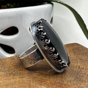 May include: A silver ring with a black oval gemstone set in a flame-shaped bezel. The ring is on a wooden surface.