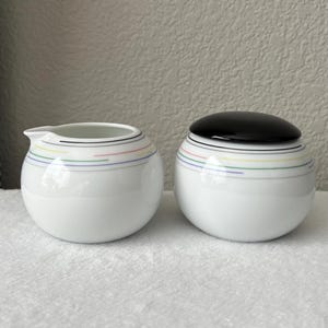 May include: A white creamer and sugar set with a black lid on the sugar bowl. Both pieces have a colorful stripe design around the middle.