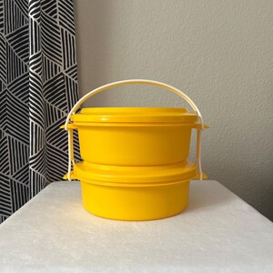 May include: Two bright yellow, round food storage containers stacked on top of each other. The containers have matching lids and a white handle for carrying. The containers are made of plastic.