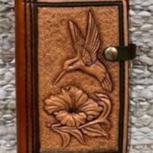 Puede incluir: Un diario de cuero marrón con un diseño tallado de un colibrí sobrevolando una flor. El diario tiene una correa de cuero marrón con un cierre de botón metálico.