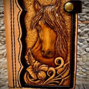 May include: A brown leather notebook cover with a carved horse head design and floral accents. The cover has a black strap with a gold button closure.