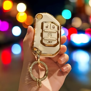 Op de afbeelding: Witte en gouden autosleutel met een gouden ring sleutelhanger. De sleutel heeft vier knoppen en een gouden logo. De sleutel wordt in een hand gehouden tegen een wazige achtergrond van kleurrijke lichten.