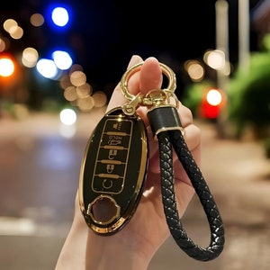 Op de afbeelding: Zwarte en gouden autosleutelhoes met een gevlochten zwart leren bandje en goudkleurige hardware. De sleutelhanger heeft een glanzende afwerking en is voorzien van sleutelsymbolen. Het CarKeysShop-logo staat rechtsboven.