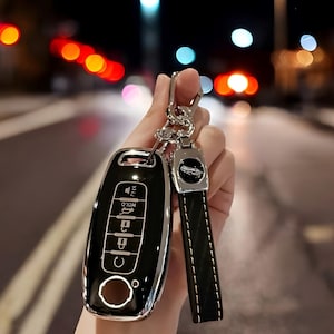 May include: A black car key fob with a silver trim and buttons labeled "Hold" and "C". The key fob is attached to a brown leather keychain with a silver car logo charm.