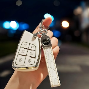 May include: White car key fob with a silver trim and multiple buttons. The key fob is attached to a white leather keychain with a silver charm.