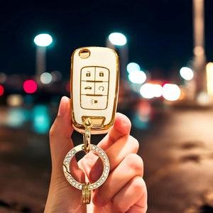 May include: A gold and white car key fob cover with a key ring. The key fob has buttons for locking, unlocking, and other functions. The key ring is adorned with sparkling crystals. The text "CarkeysShop" is in the upper right corner.