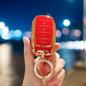 Op de afbeelding: Een rode en gouden autosleutel met een sleutelhanger met diamanten. De sleutel heeft vier knoppen: ontgrendelen, vergrendelen, kofferbak en paniek.