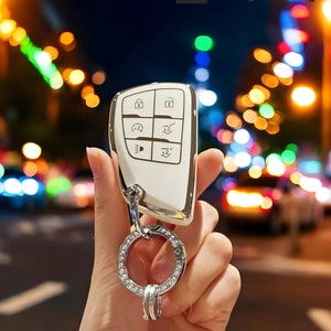 May include: A white car key fob with a silver metal ring and a silver metal key chain. The key fob has six buttons with various symbols, including a lock, unlock, boot, and panic button. The key fob is held in a hand against a blurry background of city lights.
