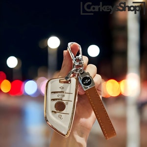 Op de afbeelding: Witte autosleutel met gouden accenten en een bruine leren sleutelhanger met een zilveren embleem. De sleutel heeft een diamantvorm en vier knoppen. De sleutelhanger heeft een zilveren embleem met een gevleugeld logo.