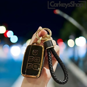 May include: A black and gold car key with a black leather braided keychain. The key has multiple buttons and a gold trim.