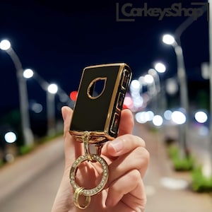 May include: A black and gold car key fob with a diamond-encrusted keychain. The key fob has a black face with a gold trim and a small keyhole cutout. The keychain is a gold ring with a circle of diamonds.