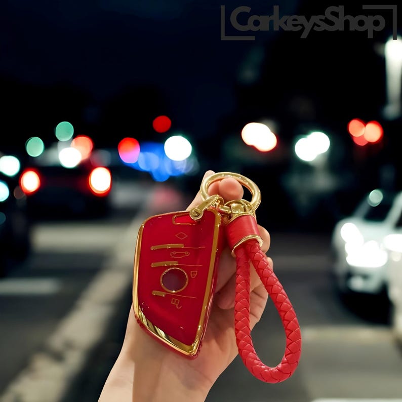 Pu&ograve; includere: Un portachiavi per auto rosso con un bordo dorato e un cinturino in pelle intrecciata rosso. Il portachiavi ha un pulsante a forma di diamante e diversi altri pulsanti.