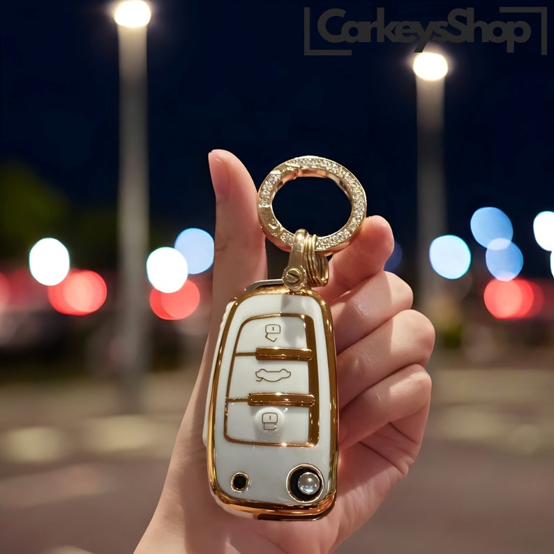 May include: A white and gold car key fob with a gold key ring. The fob has a white body with gold trim and buttons. The key ring is adorned with small, clear crystals. The background is blurred with bokeh lights.