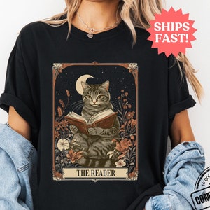 May include: A black t-shirt featuring a vintage-style illustration of a cat reading a book. The illustration is surrounded by flowers and a crescent moon. The text "THE READER" is printed below the cat.