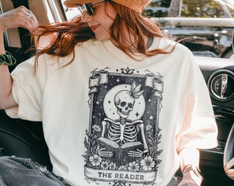 May include: A white t-shirt featuring a black and white illustration of a skeleton reading a book. The illustration is surrounded by flowers and vines. The text "THE READER" is printed below the illustration.