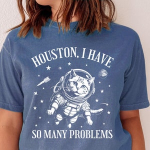 May include: A blue jean t-shirt with a white graphic of a cat wearing an astronaut suit and the text "Houston, I have so many problems".