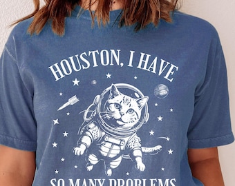 May include: A blue jean t-shirt with a white graphic of a cat wearing an astronaut suit and the text "Houston, I have so many problems".