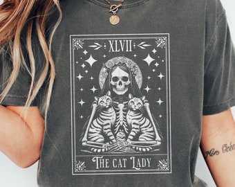 May include: A pepper-coloured t-shirt with a tarot card design. The graphic shows a skeleton with a floral crown, two cats, and the text "The Cat Lady." The design includes the Roman numeral XLVII and star accents.