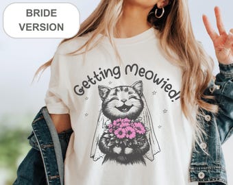 May include: White t-shirt with a graphic of a cat wearing a veil and holding a bouquet of flowers. The text "Getting Meowied!" is printed on the shirt.