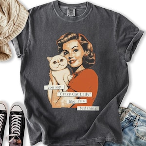 May include: Dark gray t-shirt featuring a vintage-style illustration of a woman holding a white cat. The woman is wearing an orange top. Text on the shirt reads "you say 'Crazy Cat Lady' like it's a bad thing."