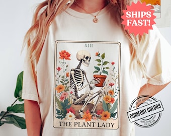 May include: A white t-shirt with a tarot card design featuring a skeleton holding a potted plant. The card is titled "The Plant Lady" and has the Roman numeral XIII at the top. The shirt has a tag that says "Comfort Colors" and "100% Ring-Spun US Cotton".