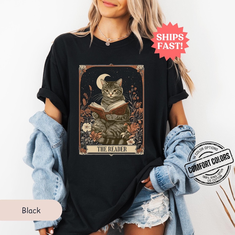 May include: A black t-shirt featuring a vintage-style illustration of a cat reading a book. The illustration is surrounded by flowers and a crescent moon. The text "THE READER" is printed below the cat.