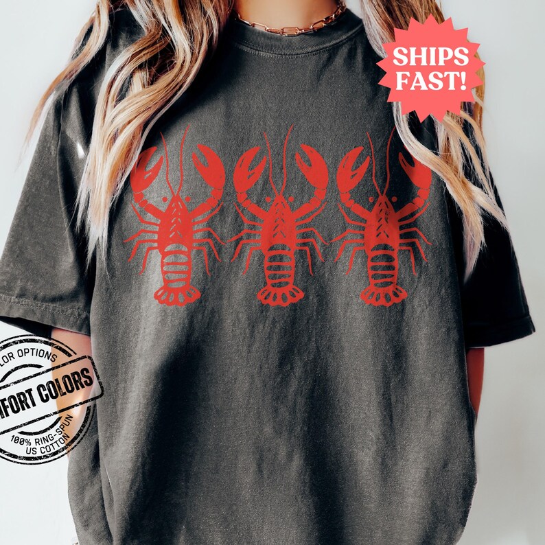 Puede incluir: Una camiseta gris oscura con un gr&aacute;fico rojo de tres langostas. Las langostas est&aacute;n mirando hacia adelante con las garras levantadas.