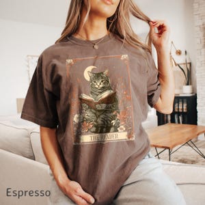 May include: A brown t-shirt featuring a vintage illustration of a cat reading a book. The illustration is surrounded by flowers and has a crescent moon in the background. The text "The Reader" is printed below the illustration.