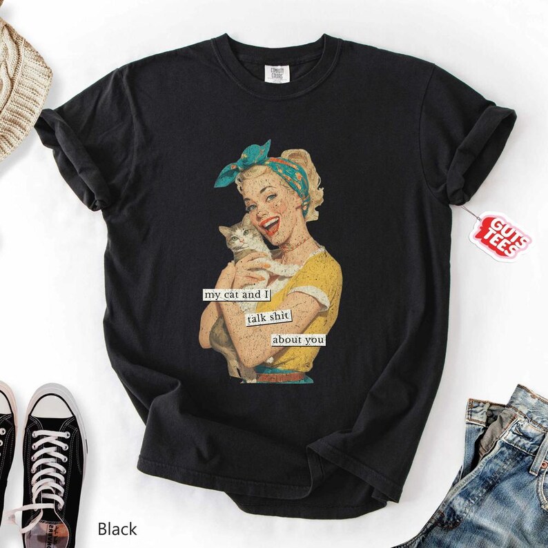 Puede incluir: Camiseta negra con una ilustraci&oacute;n retro de una mujer sosteniendo un gato. La mujer lleva una blusa amarilla y un pa&ntilde;uelo azul. El texto en la camiseta dice "my cat and I talk shit about you."