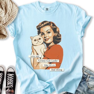 May include: Light blue t-shirt featuring a retro illustration of a woman holding a white cat. The text reads "you say 'Crazy Cat Lady' like it's a bad thing". The shirt has a crew neck and short sleeves. The shirt is made of a soft material.