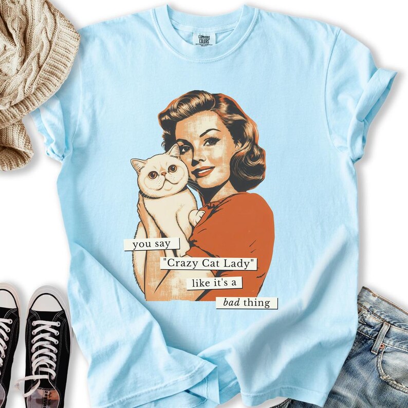 May include: Light blue t-shirt featuring a retro illustration of a woman holding a white cat. The text reads "you say 'Crazy Cat Lady' like it's a bad thing". The shirt has a crew neck and short sleeves. The shirt is made of a soft material.