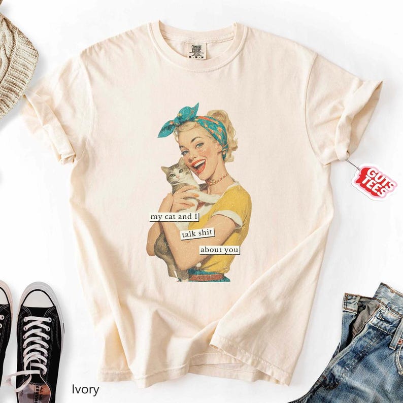 Puede incluir: Camiseta color marfil con una ilustraci&oacute;n retro de una mujer sosteniendo un gato. La mujer lleva un top amarillo y un pa&ntilde;uelo azul. El texto en la camiseta dice "my cat and I talk shit about you."