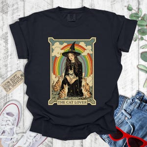 May include: A black t-shirt with a vintage-style illustration of a witch with four cats. The illustration is surrounded by a rainbow and the text "The Cat Lover".