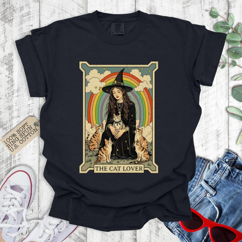 May include: A black t-shirt with a vintage-style illustration of a witch with four cats. The illustration is surrounded by a rainbow and the text "The Cat Lover".