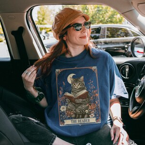 May include: A blue t-shirt featuring a graphic of a cat reading a book beneath a crescent moon. The text "The Reader" is printed below the image. The person wearing the shirt is seated in a car.