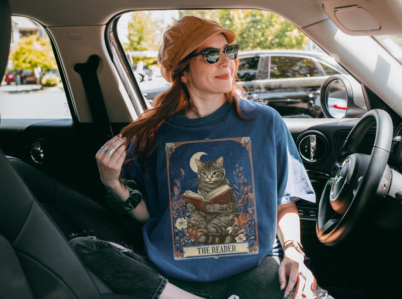 May include: A blue t-shirt featuring a graphic of a cat reading a book beneath a crescent moon. The text "The Reader" is printed below the image. The person wearing the shirt is seated in a car.