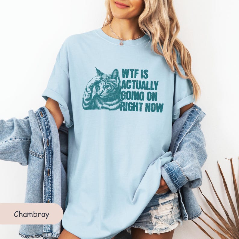 May include: A light blue t-shirt with a graphic of a cat with the text "WTF IS ACTUALLY GOING ON RIGHT NOW" printed in teal.