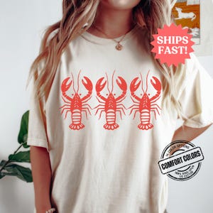 Puede incluir: Una camiseta blanca con un gr&aacute;fico rojo de tres langostas. El texto "SHIPS FAST!" est&aacute; en una estrella roja en la esquina superior derecha. El texto "14 COLOR OPTIONS" est&aacute; en un c&iacute;rculo rojo con un contorno blanco. El texto "COMFORT COLORS" est&aacute; en un c&iacute;rculo blanco con un contorno rojo. El texto "100% RING-SPUN US COTTON" est&aacute; en un c&iacute;rculo blanco con un contorno rojo.