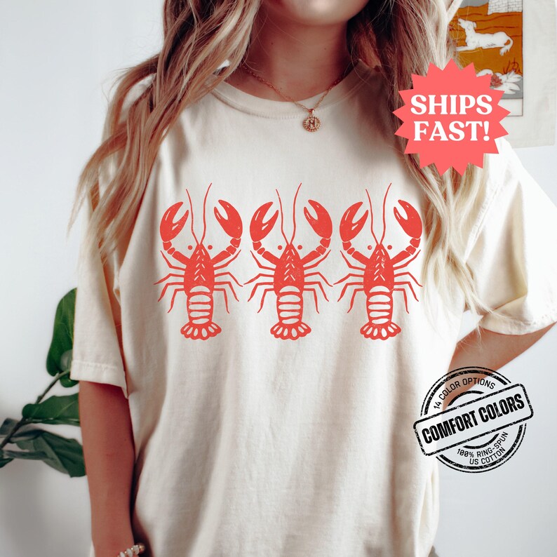 Puede incluir: Una camiseta blanca con un gr&aacute;fico rojo de tres langostas. El texto "SHIPS FAST!" est&aacute; en una estrella roja en la esquina superior derecha. El texto "14 COLOR OPTIONS" est&aacute; en un c&iacute;rculo rojo con un contorno blanco. El texto "COMFORT COLORS" est&aacute; en un c&iacute;rculo blanco con un contorno rojo. El texto "100% RING-SPUN US COTTON" est&aacute; en un c&iacute;rculo blanco con un contorno rojo.