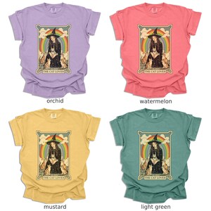 May include: Four t-shirts in different colors, each featuring a vintage-style illustration of a woman with a cat. The illustration is surrounded by a rainbow and the text "The Cat Lover". The t-shirts are available in orchid, watermelon, mustard, and light green.