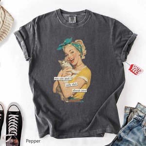 Puede incluir: Camiseta gris oscuro con una ilustraci&oacute;n vintage de una mujer sosteniendo un gato. La mujer lleva una camiseta amarilla y un pa&ntilde;uelo azul. El texto en la camiseta dice "my cat and I talk shit about you."
