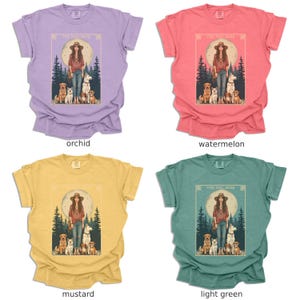 May include: Four different coloured t-shirts with a graphic design of a woman with five dogs in a forest setting. The text on the shirt reads "The Dog Mom". The t-shirts are in the colours orchid, watermelon, mustard, and light green.