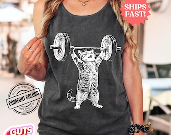 May include: Dark gray tank top featuring a white graphic of a cat lifting a barbell. The tank top has a scoop neck and is made of a soft, comfortable material. The text "GUTS TEES" is visible. The tank top is available in 14 color options.
