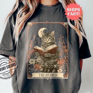 May include: A vintage-style tarot card illustration featuring a grey tabby cat reading a book under a crescent moon. The card is surrounded by flowers and the text "THE READER" is printed at the bottom.
