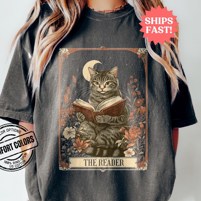 May include: A vintage-style tarot card illustration featuring a grey tabby cat reading a book under a crescent moon. The card is surrounded by flowers and the text "THE READER" is printed at the bottom.