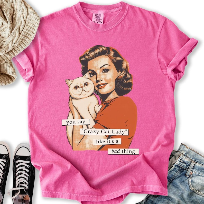 May include: Pink t-shirt featuring a vintage-style illustration of a woman holding a white cat. The text on the shirt reads "you say 'Crazy Cat Lady' like it's a bad thing."