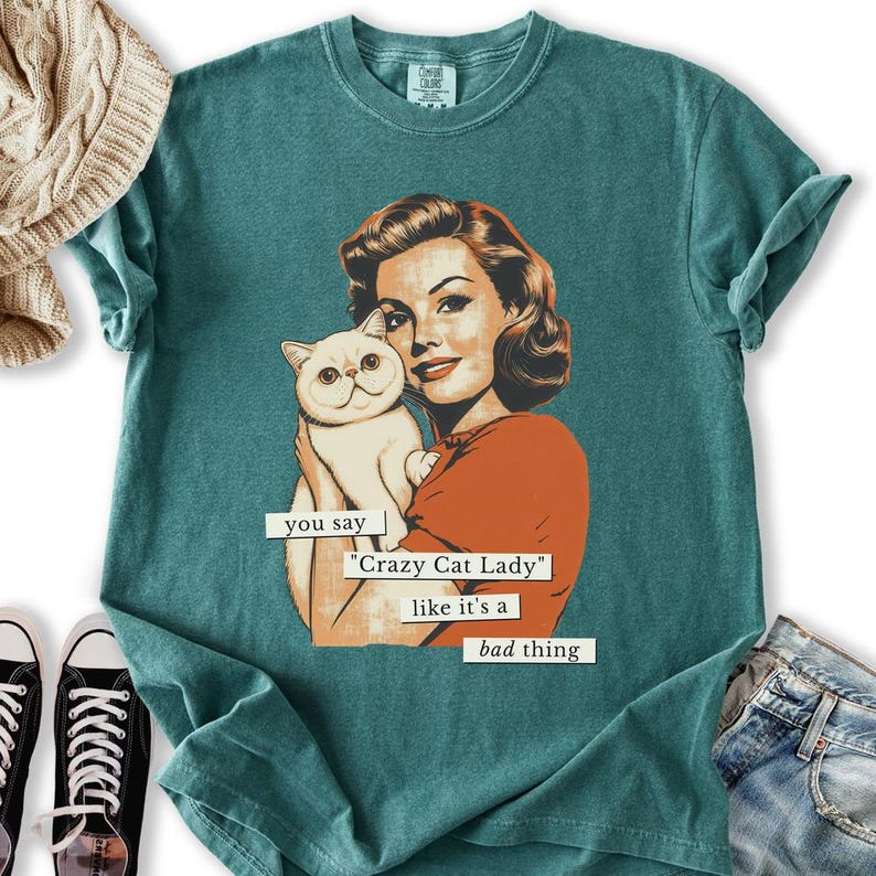 May include: Teal-colored t-shirt featuring a vintage-style illustration of a woman holding a white cat. The text on the shirt reads "you say 'Crazy Cat Lady' like it's a bad thing."