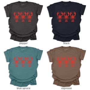 Puede incluir: Cuatro ilustraciones de langostas rojas en una camiseta. La camiseta est&aacute; disponible en cinco colores: pimienta, negro, abeto azul, espresso y natural.