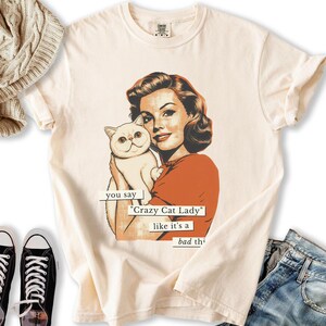 May include: Cream-colored t-shirt featuring a retro illustration of a woman holding a white cat. The text reads "you say 'Crazy Cat Lady' like it's a bad thing." The woman is wearing an orange top. The shirt has a classic, vintage aesthetic.