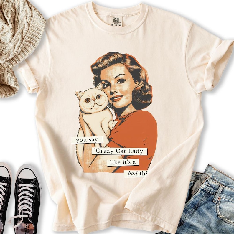 May include: Cream-colored t-shirt featuring a retro illustration of a woman holding a white cat. The text reads "you say 'Crazy Cat Lady' like it's a bad thing." The woman is wearing an orange top. The shirt has a classic, vintage aesthetic.